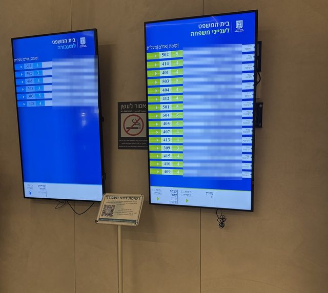לוח הדיונים האלקטרוני בכניסה — בית המשפט לתעבורה, עם מספר האולם והקומה לכל דיון, ולצידו שלט QR לרשימת דיוני התעבורה